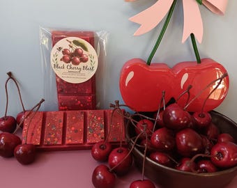 May include: A display featuring cherry-themed decor and products. A package of wax melts labeled "Black Cherry Blast" sits next to a bar of wax melts. A decorative cherry bow and a bowl of cherries complete the arrangement. The color scheme is red, pink, and green.