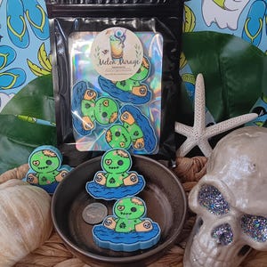 May include: A black bag with several zombie-themed stickers, featuring a green zombie character floating on blue water. The stickers are in a small bowl with a coin. The background includes a skull, starfish, and a beach-themed pattern.