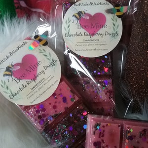 May include: Two wax melts in clear packaging with a pink and purple glitter design. The wax melts are labeled "Bee Mine Chocolate Raspberry Drizzle" with the ingredients listed below. The wax melts are packaged in a clear plastic bag with a white fuzzy background.