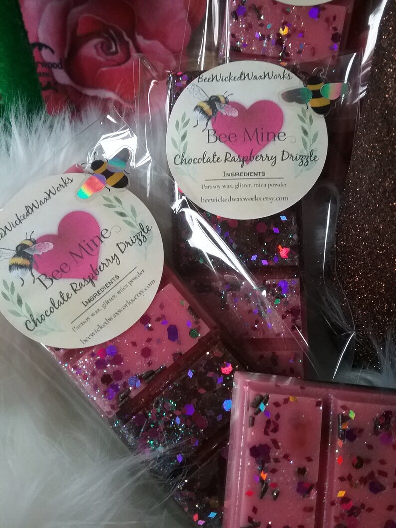 May include: Two wax melts in clear packaging with a pink and purple glitter design. The wax melts are labeled "Bee Mine Chocolate Raspberry Drizzle" with the ingredients listed below. The wax melts are packaged in a clear plastic bag with a white fuzzy background.