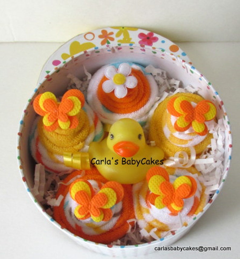 May include: A yellow rubber ducky in a gift box with four rolled up towels decorated with orange and yellow butterflies. The towels are arranged in a circle around the ducky. The box is decorated with a colorful floral pattern.