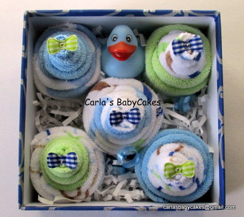 May include: A blue and white gift box filled with six baby shower favors. Each favor is a stack of blue and white towels with a green bow. The box also includes a blue rubber duck.