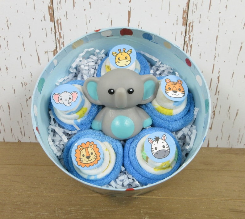 May include: A blue and white polka dot tin filled with blue and white baby washcloths rolled into a flower shape. Each washcloth has a different animal face on it: a giraffe, a tiger, a zebra, an elephant, and a lion. A gray elephant bath toy is in the center of the arrangement.