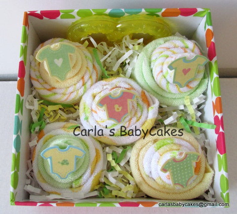 May include: A gift box filled with four yellow and green baby washcloths rolled into the shape of cupcakes. Each cupcake is topped with a small felt baby onesie. The box is lined with white shredded paper and has a colorful polka dot pattern.