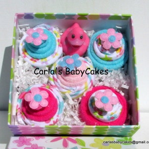May include: A pink and blue baby shower gift set in a box with a pink rubber ducky in the center. The gift set includes four diaper cakes, each with a different color and pattern.