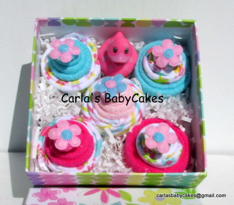 May include: A pink and blue baby shower gift set in a box with a pink rubber ducky in the center. The gift set includes four diaper cakes, each with a different color and pattern.