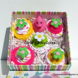 May include: A pink and yellow gift box filled with four towel cupcakes. Each cupcake is decorated with a green flower and pink and white polka dots. The box is lined with white tissue paper and has a pink and white striped lid. The text "Carla's BabyCakes" is printed on the box.