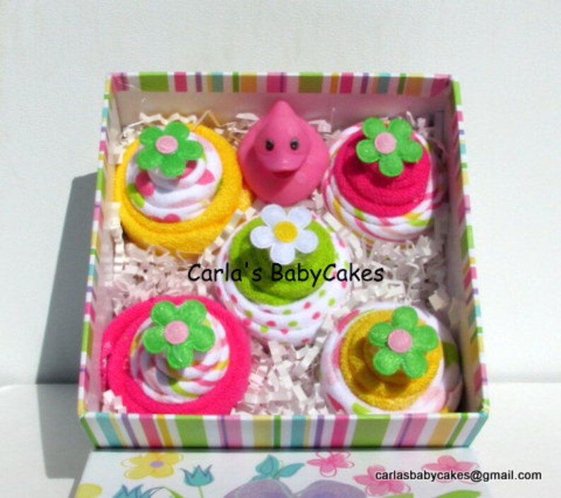 May include: A pink and yellow gift box filled with four towel cupcakes. Each cupcake is decorated with a green flower and pink and white polka dots. The box is lined with white tissue paper and has a pink and white striped lid. The text "Carla's BabyCakes" is printed on the box.