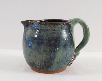 May include: A small, green and brown ceramic pitcher with a rounded body and a handle. The pitcher has a glazed finish.