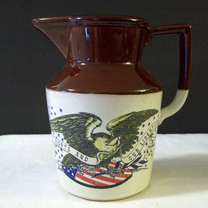 May include: A brown and white ceramic pitcher with a handle. The pitcher features an eagle design with the words "OUR LIBERTIES AND RIGHTS" and a red, white, and blue flag. The top and handle are brown.