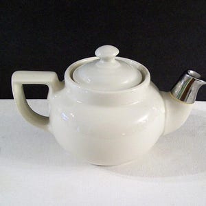 May include: A white ceramic teapot with a rounded body, a handle, and a lid with a small knob. The spout has a silver metal finish. The teapot is on a white surface with a black background.
