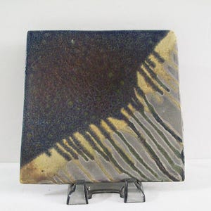 May include: Square ceramic tile with a unique glaze. The tile features a dark blue and brown color scheme with streaks of yellow and gray. The tile is displayed on a clear stand.