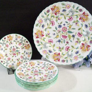 May include: A collection of white ceramic plates with a floral pattern in pink, yellow, and blue. The plates have a scalloped edge with a green trim. The set includes dinner plates and smaller plates, perfect for a formal dining setting.