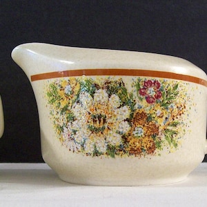 May include: A set of two ceramic creamers with a floral design. The creamers are white with a brown rim and a floral pattern in shades of yellow, red, and green.