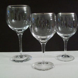 May include: Three clear glass wine glasses with rounded bowls and slender stems. Each glass features a thin, reflective silver metallic rim around the top edge. The glasses are arranged in a row, showcasing their transparent construction and polished finish.