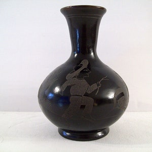 May include: A black ceramic vase with a wide base and a narrow neck. The vase is decorated with a black and white silhouette of a person wearing a hat and holding a stick.