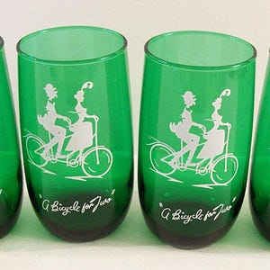 May include: Set of four green glass tumblers with a white silhouette design of a couple riding a bicycle. Each glass has the text "A Bicycle for Two" printed on the bottom.