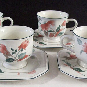 May include: Set of four floral teacups and saucers. The cups and saucers are white with a green border and a floral design in shades of pink, green, and brown.