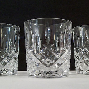 May include: Three clear crystal drinking glasses with diamond-cut patterns. The glasses are cylindrical with a slightly flared rim. The crystal reflects light, creating a sparkling effect. The glasses are arranged on a white surface against a black background.