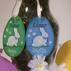 May include: Two wooden Easter egg ornaments with painted bunnies and flowers. The green ornament says "My 1st Easter" and "Annie 2016". The blue ornament says "My 1st Easter" and "Justin 2016".