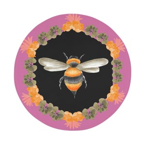 May include: A pink round plate with a black center featuring a detailed illustration of a bee with orange and black stripes. The plate is decorated with a border of orange and green flowers.