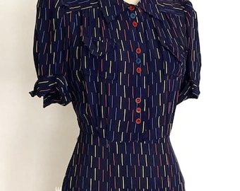 May include: A navy blue midi dress featuring a repeating pattern of vertical dashes in red, yellow, and blue. The dress has short puff sleeves, a collared neckline, two chest pockets, and a front button placket with multicolored buttons.