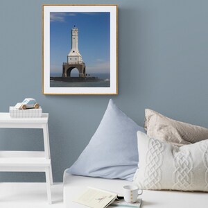 May include: A framed print of a white lighthouse with a tall tower and arched base, set against a blue sky. The artwork is displayed on a blue wall, with a white bed, pillows, and a small table in the foreground. A toy car and a book are also visible.