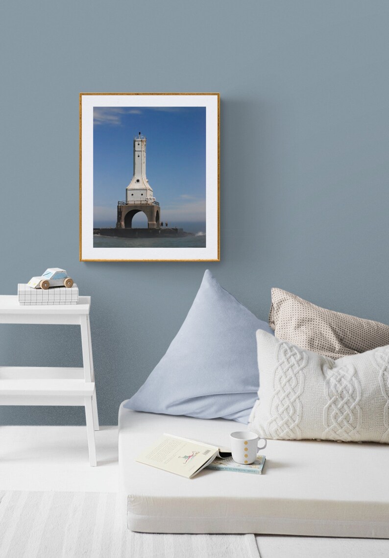May include: A framed print of a white lighthouse with a tall tower and arched base, set against a blue sky. The artwork is displayed on a blue wall, with a white bed, pillows, and a small table in the foreground. A toy car and a book are also visible.