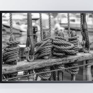 Pu&ograve; includere: Fotografia in bianco e nero incorniciata di sartiame di una nave. Spesse corde sono avvolte attorno a pali e travi di legno. L'immagine cattura la consistenza delle corde e del legno, con uno sfondo sfocato di barche e acqua.