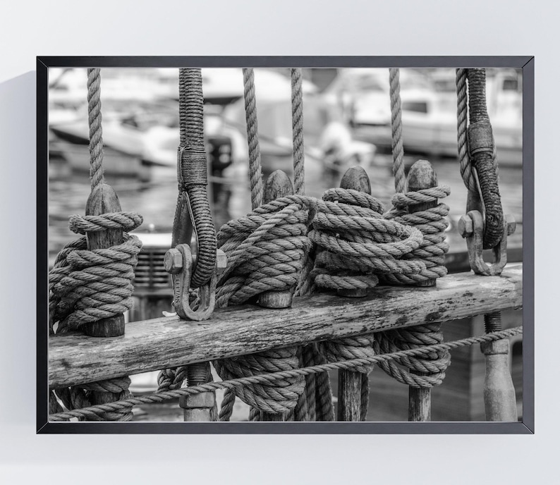 Pu&ograve; includere: Fotografia in bianco e nero incorniciata di sartiame di una nave. Spesse corde sono avvolte attorno a pali e travi di legno. L'immagine cattura la consistenza delle corde e del legno, con uno sfondo sfocato di barche e acqua.