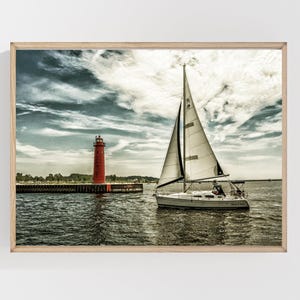 May include: A framed print of a sailboat sailing on a body of water, with a red lighthouse in the background. The sky is cloudy with shades of blue and white. The sailboat has a white sail and a dark hull.