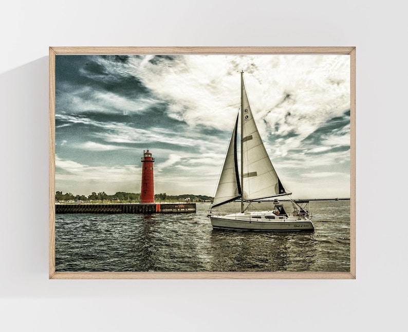 May include: A framed print of a sailboat sailing on a body of water, with a red lighthouse in the background. The sky is cloudy with shades of blue and white. The sailboat has a white sail and a dark hull.