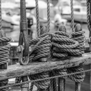 Pu&ograve; includere: Immagine in bianco e nero di una sezione di una barca a vela. Spesse corde sono avvolte attorno a pali di legno e fissate con ferramenta metallica. L'immagine si concentra sugli intricati dettagli del sartiame, con uno sfondo sfocato di altre barche.