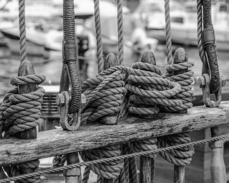 Pu&ograve; includere: Immagine in bianco e nero di una sezione di una barca a vela. Spesse corde sono avvolte attorno a pali di legno e fissate con ferramenta metallica. L'immagine si concentra sugli intricati dettagli del sartiame, con uno sfondo sfocato di altre barche.