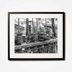 Pu&ograve; includere: Fotografia in bianco e nero incorniciata di un'attrezzatura navale. L'immagine si concentra sul ponte di legno e sulle corde, con bobine avvolte strettamente e corde verticali. Lo sfondo &egrave; sfocato, suggerendo un porto o acque libere.