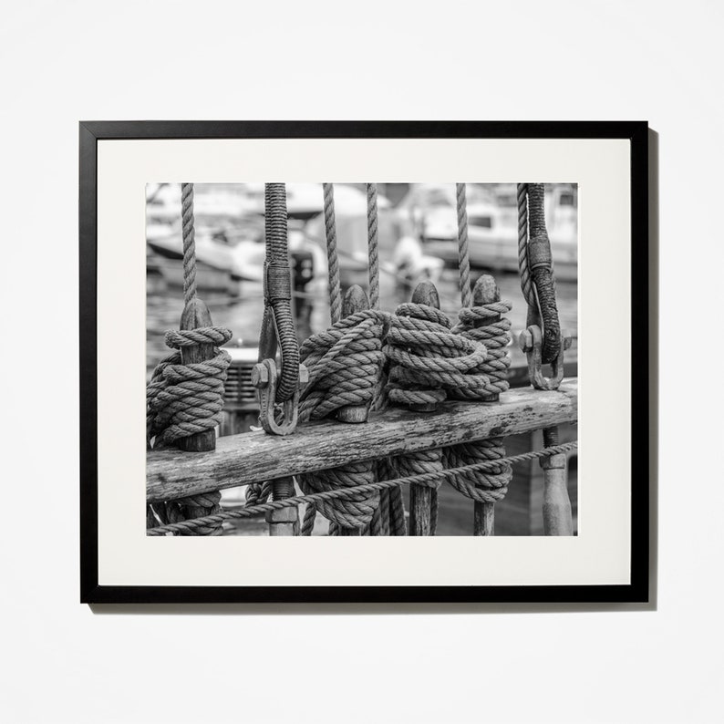 Pu&ograve; includere: Fotografia in bianco e nero incorniciata di un'attrezzatura navale. L'immagine si concentra sul ponte di legno e sulle corde, con bobine avvolte strettamente e corde verticali. Lo sfondo &egrave; sfocato, suggerendo un porto o acque libere.