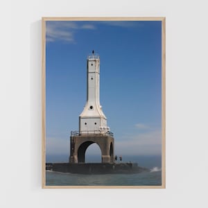 May include: A framed print of a white lighthouse against a bright blue sky. The lighthouse has a tall, rectangular tower with a black top and is connected to a concrete pier. The water is choppy, and the overall scene is bright and sunny.