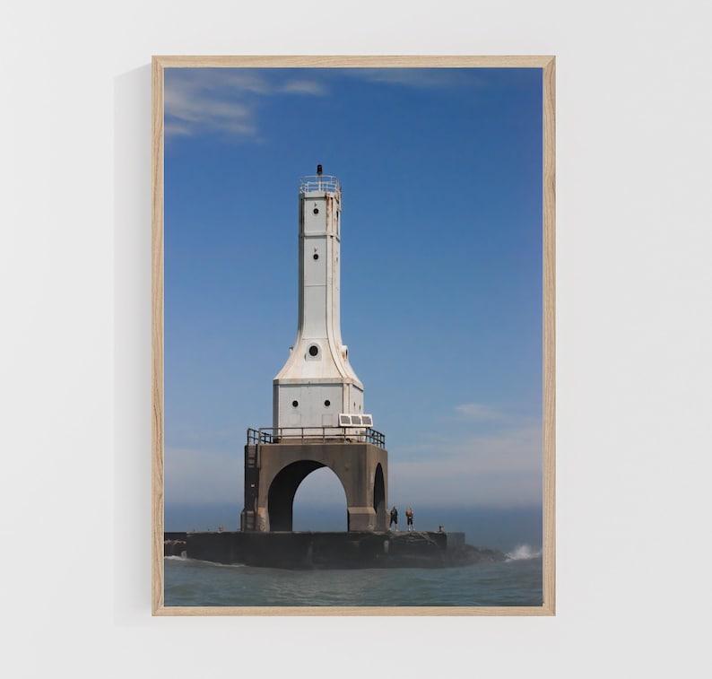 May include: A framed print of a white lighthouse against a bright blue sky. The lighthouse has a tall, rectangular tower with a black top and is connected to a concrete pier. The water is choppy, and the overall scene is bright and sunny.