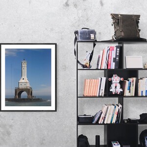 May include: A framed photograph of a white lighthouse with a black frame hangs on a gray wall. A black bookshelf is to the right, holding books, a backpack, a camera bag, and decorative items. The lighthouse is on a stone base.