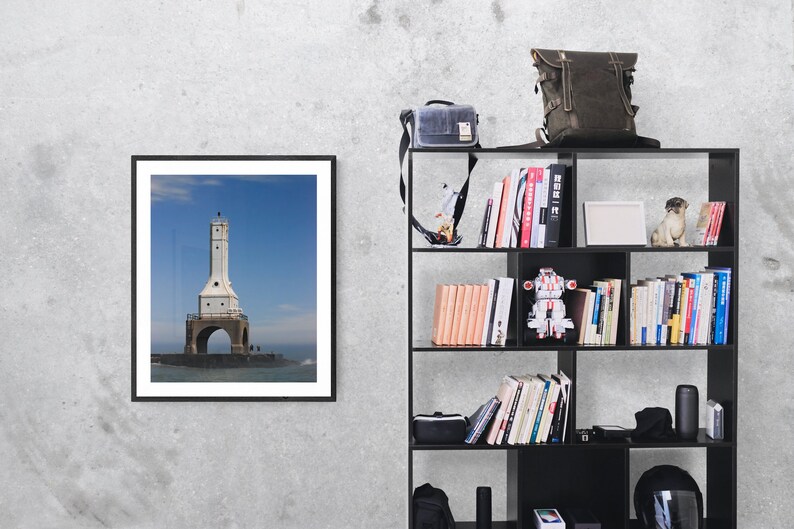 May include: A framed photograph of a white lighthouse with a black frame hangs on a gray wall. A black bookshelf is to the right, holding books, a backpack, a camera bag, and decorative items. The lighthouse is on a stone base.