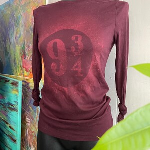 Könnte beinhalten: Ein weinrotes Langarmshirt mit der Aufschrift 93 4 auf der Vorderseite. Die Ärmel sind an den Handgelenken geknotet. Das Shirt wird auf einer schwarzen Schaufensterpuppe präsentiert.