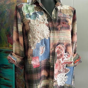 Peut inclure: Une chemise à manches longues boutonnée avec un motif à carreaux dans des tons terreux. Le vêtement est orné de divers empiècements en tissu, incluant des imprimés floraux, du denim, de la dentelle et un motif léopard, créant un aspect texturé.