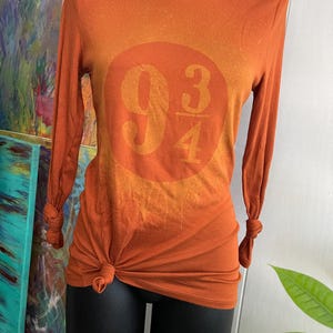 Könnte beinhalten: Orangefarbenes Langarmshirt mit einem runden Aufdruck mit den Zahlen "9 3/4" in einem helleren Orangeton. Das Shirt ist an der Taille und die Ärmel an den Handgelenken geknotet. Das Shirt wird auf einer schwarzen Schaufensterpuppe präsentiert.
