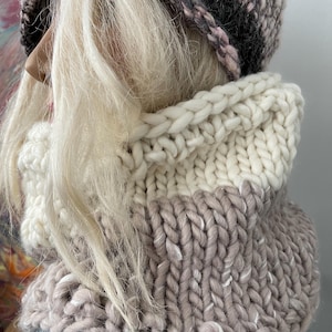 May include: A white and gray knitted cowl neck scarf with a chunky knit texture. The scarf is worn with a gray and pink knitted hat.
