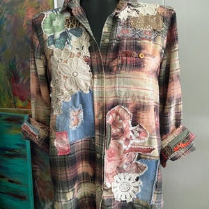 May include: A long-sleeve button-down shirt featuring a plaid pattern in muted tones of brown, orange, and green. The garment is embellished with various fabric patches, including floral prints, denim, lace doilies, and textured textile scraps.