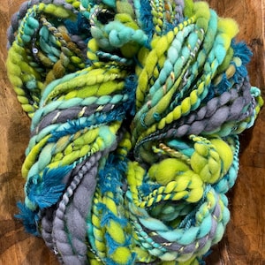 Puede incluir: Una madeja de hilo artístico con una mezcla de fibras verde lima, azul cerceta, turquesa y gris carbón. El hilo presenta diversas texturas, incluyendo mechas gruesas, hebras retorcidas, fibras esponjosas y finos hilos metálicos entrelazados.