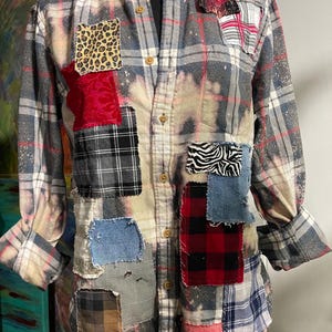 Peut inclure: Une chemise à carreaux boutonnée à manches longues avec un motif patchwork. La chemise présente un motif à carreaux gris et rouge, agrémenté de divers patchs en tissu à imprimé animal, en denim et à carreaux. La chemise a un aspect blanchi et vieilli.