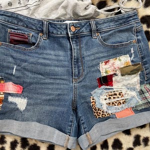 May include: Blue denim shorts with rolled cuffs and various fabric patches. Patches feature floral, plaid, and animal print patterns. The shorts have a button closure and classic five-pocket design.