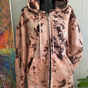 May include: A peach-colored zip-up hoodie with a brown, tie-dye-like pattern. The hoodie features a drawstring hood, a full-length zipper, and two front pockets. The garment is displayed on a black mannequin.