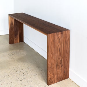 May include: A long, rectangular walnut wood console table with a simple, modern design. The table has a wide, flat top and two vertical supports on either end.